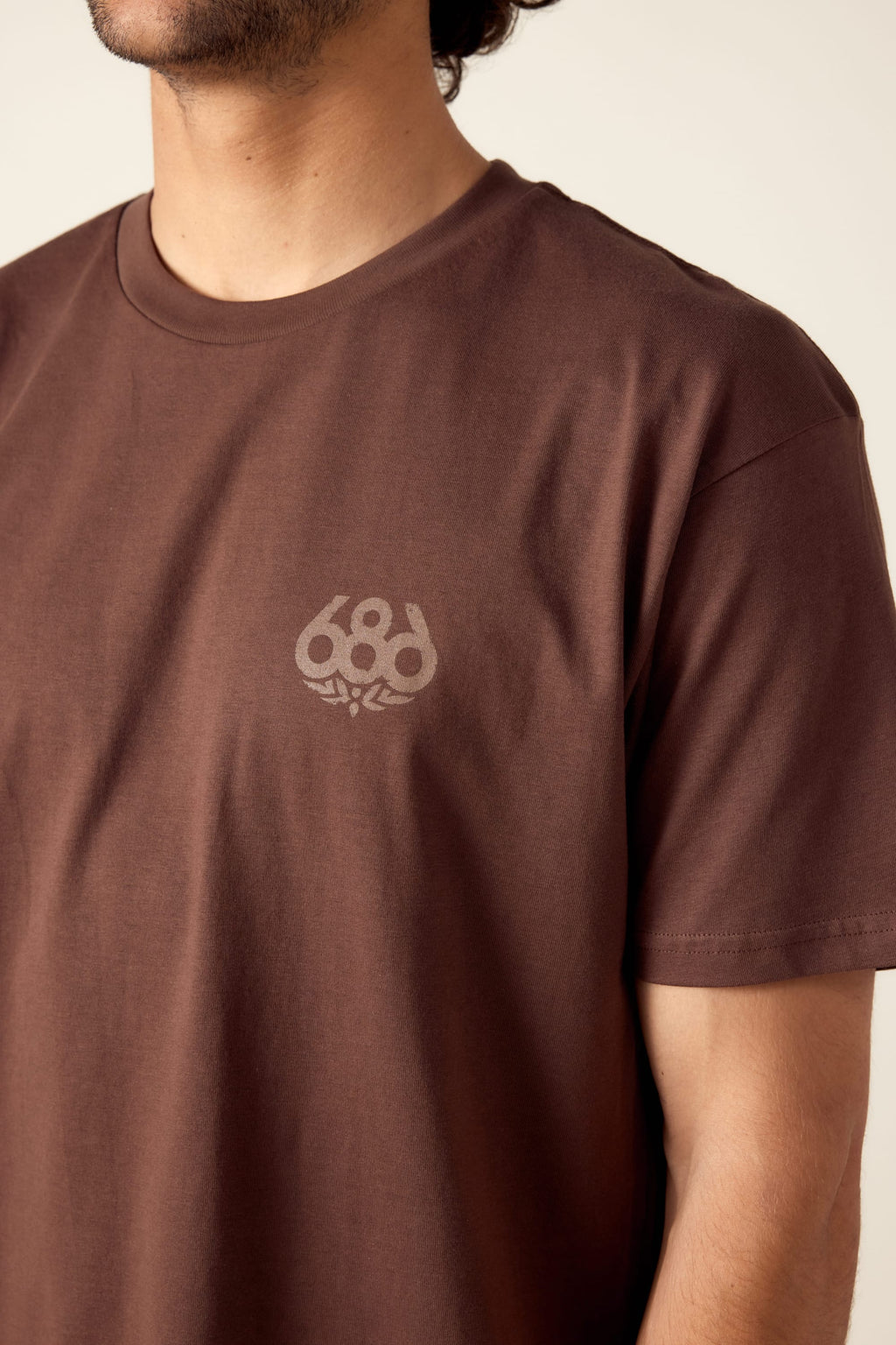 A person wearing the 686 Knockout Imprint Authentic S/S Tee, a brown men’s cotton t-shirt with a small 686 logo on the left chest, is shown from upper torso to part of face against a neutral beige background.