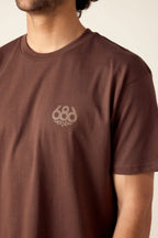 A person wearing the 686 Knockout Imprint Authentic S/S Tee, a brown men’s cotton t-shirt with a small 686 logo on the left chest, is shown from upper torso to part of face against a neutral beige background.