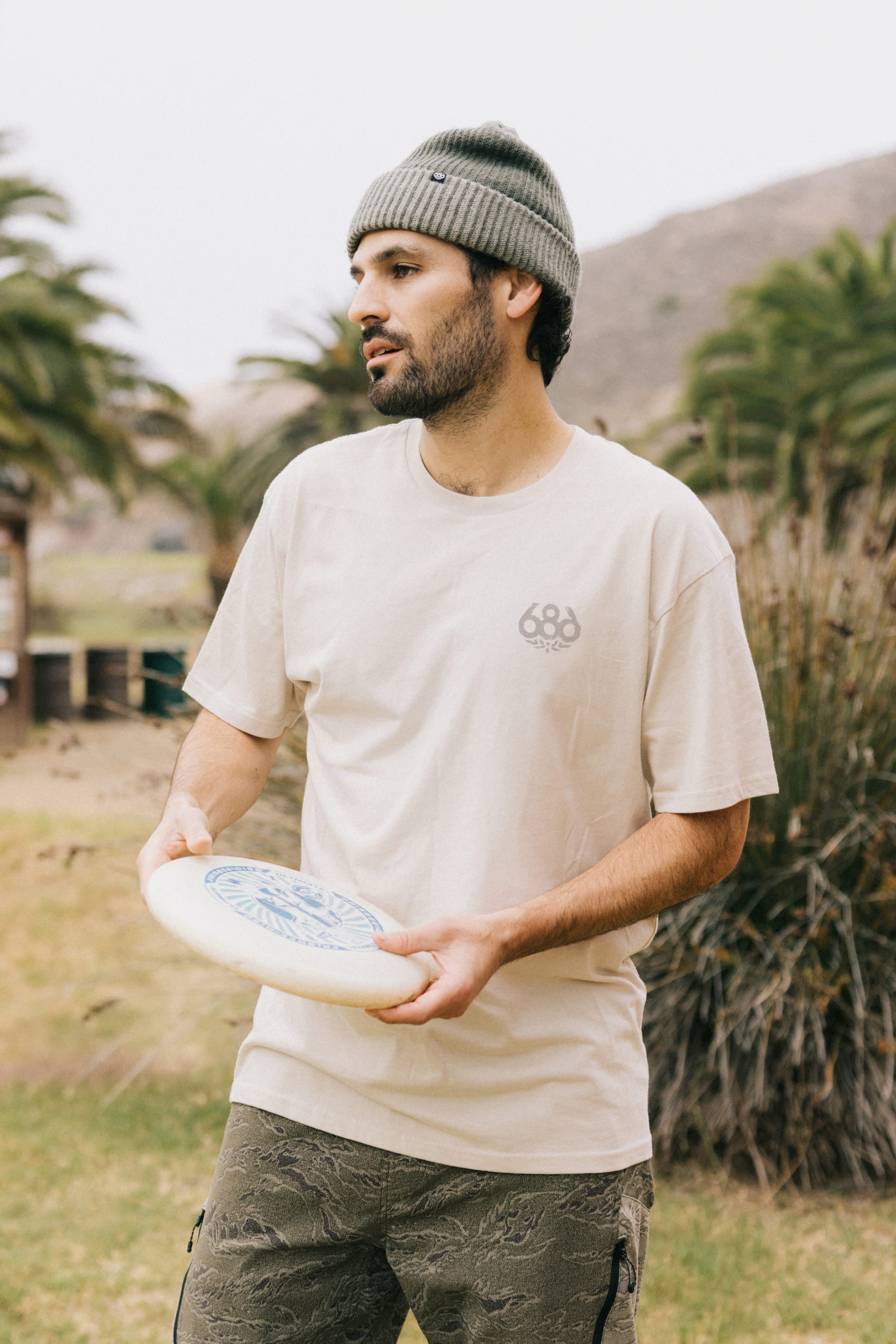 A man wearing a gray beanie and the 686 Knockout Imprint Authentic S/S Tee by 686 holds a frisbee outdoors, with palm trees and hills in the background.