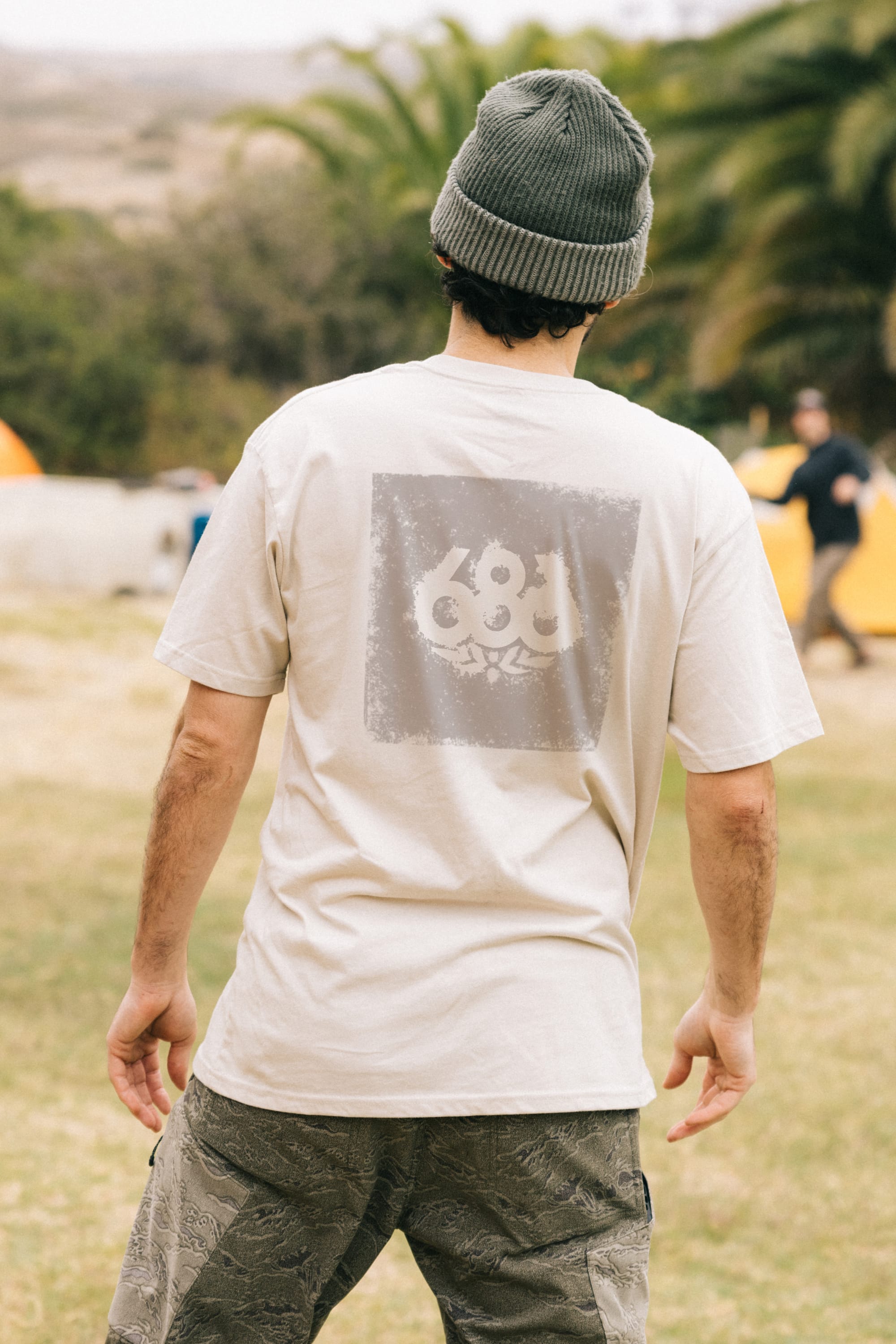 Outdoors on grass, a person in a 686 Knockout Imprint Authentic S/S Tee by 686, featuring a bold abstract back graphic, stands with palm trees and tents behind them. A beanie completes their relaxed, effortless look.