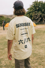 A person with wavy hair in a cap and a light-colored 686 Unwork Wear Authentic S/S Tee, featuring “SIX8SIX,” “UNWORK WEAR,” “SINCE 1992,” and Chinese characters on the back, stands on grass near orange tents and palm trees.
