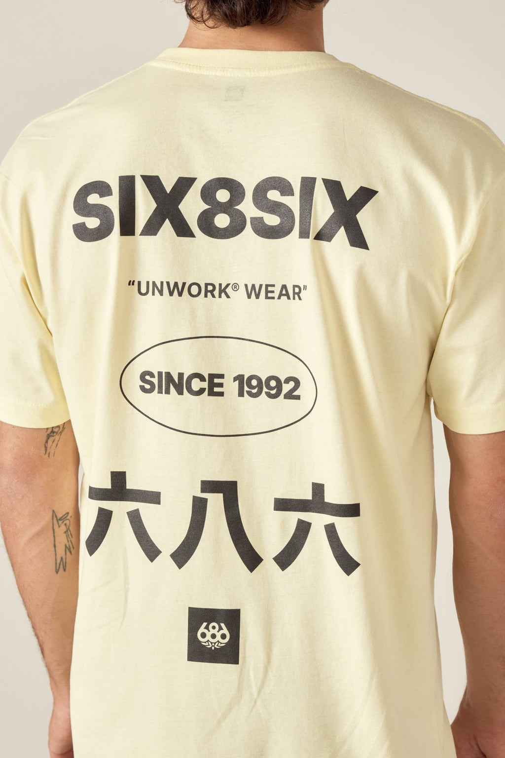 A man wears the 686 Unwork Wear Authentic S/S Tee by 686—a light yellow, 100% cotton shirt with bold black SIX8SIX, UNWORK® WEAR, SINCE 1992 text, Japanese characters, and a small circular logo on the back.