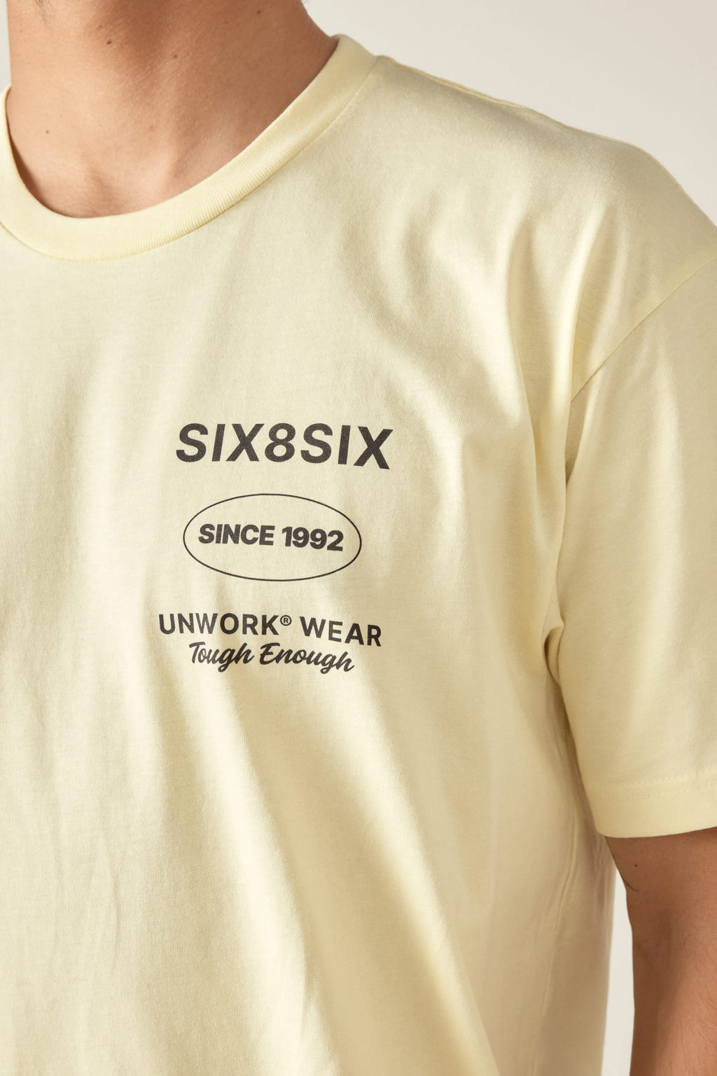 A person wears the 686 Unwork Wear Authentic S/S Tee, a pale yellow 100% cotton mens shirt with SIX8SIX, SINCE 1992, UNWORK® WEAR, Tough Enough in black on the left chest. Photo shows upper torso and part of neck. Brand: 686.