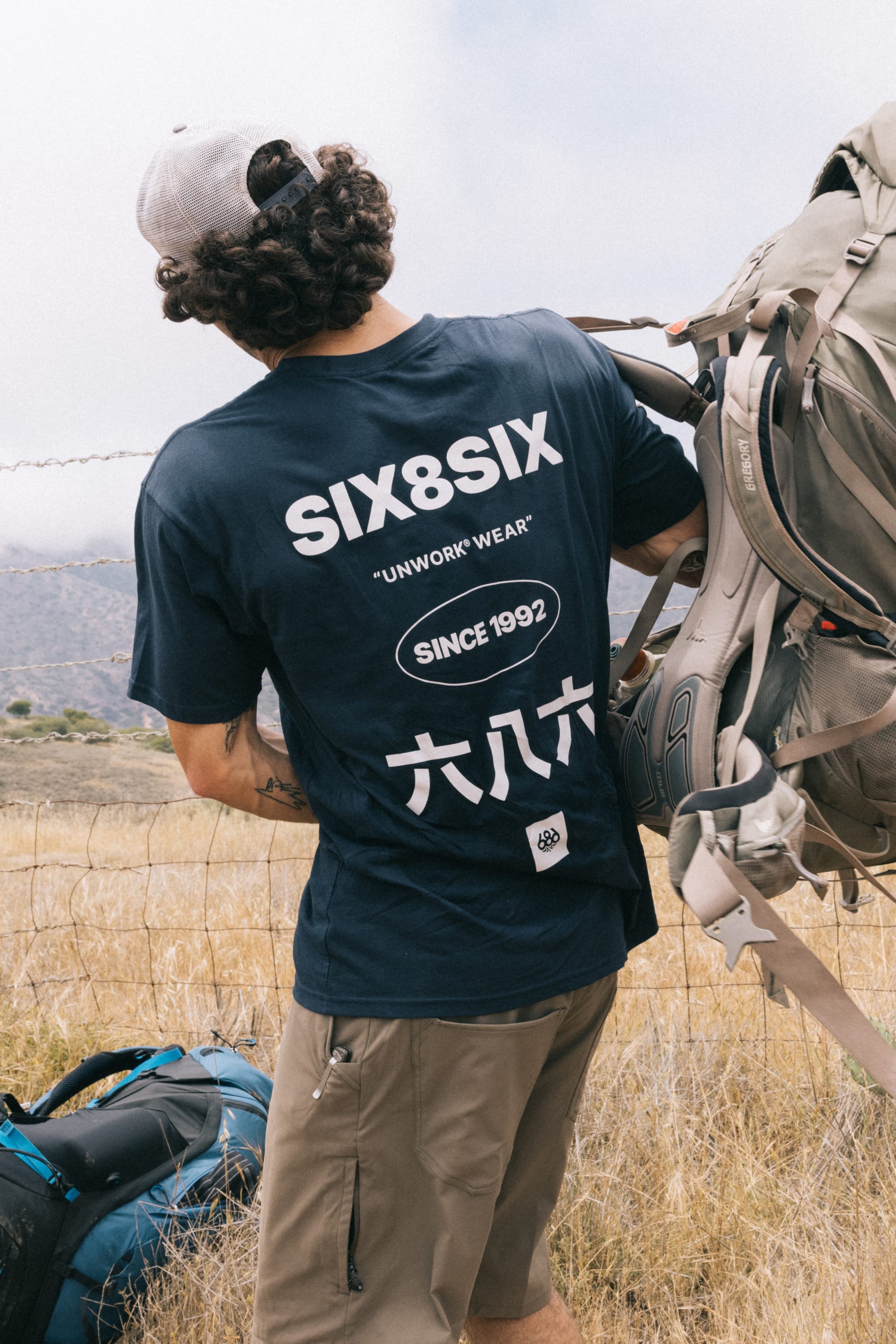 A person with curly hair, wearing a cap and a navy 686 Unwork Wear Authentic S/S Tee by 686, stands outdoors near a wire fence with a large backpack. Dry grass and hills are visible in the background.