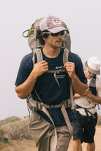 A man wearing sunglasses, a cap, and the 686 Unwork Wear Authentic S/S Tee by 686 hikes with a large backpack. Another person in a white shirt and cap follows behind on an outdoor trail.