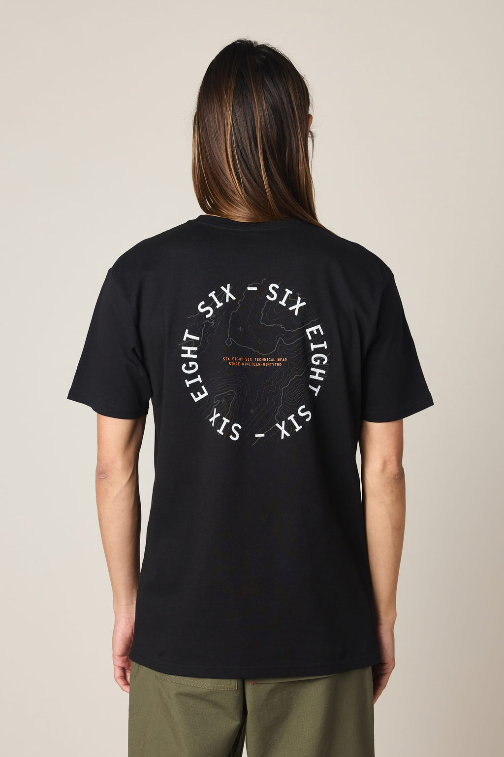 A person with long hair, seen from behind, wears the 686 Lifts Authentic SS Tee— a black 100% cotton t-shirt by 686 featuring a white “SIX EIGHT SIX” circle logo and subtle central graphic—paired with green pants.