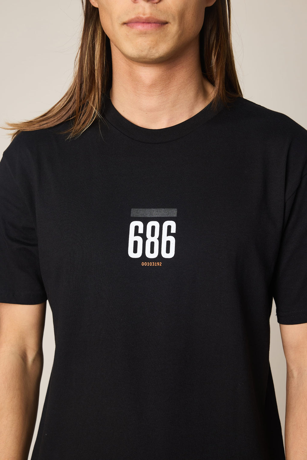 A person with long brown hair wears the black 686 Lifts Authentic SS Tee by 686, featuring "686" and "00101992" printed in white and orange on the chest. The image shows their upper torso and part of their face.