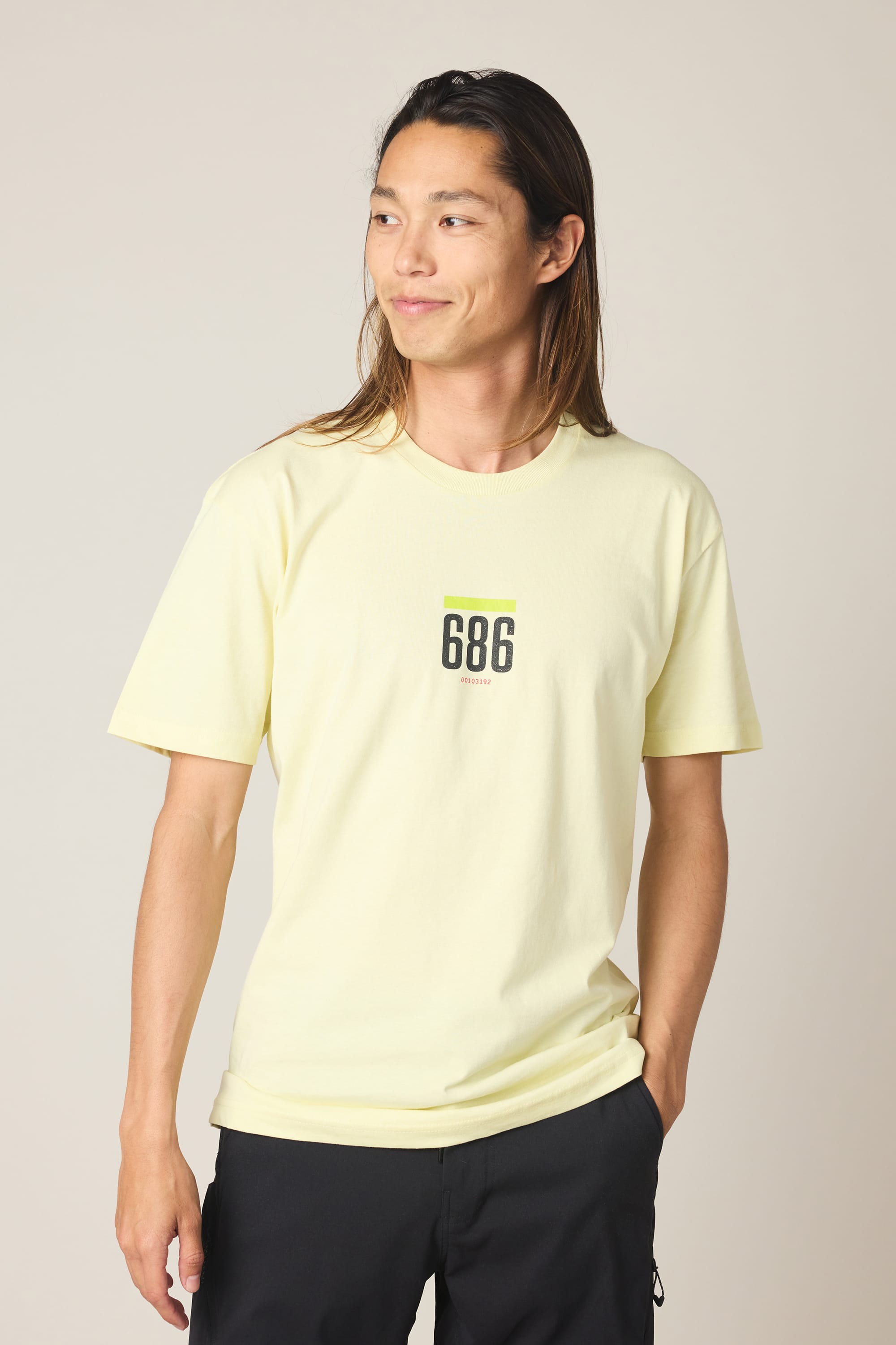 A person with long brown hair stands against a plain background, smiling slightly and looking to the side with one hand in their pocket, wearing the 686 Lifts Authentic SS Tee by 686 in light yellow.