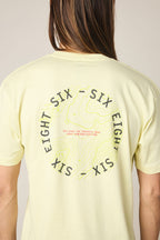 A person wearing the 686 Lifts Authentic SS Tee, a men's 100% cotton t-shirt in light yellow by 686, with "SIX EIGHT SIX" and "TECHNICAL TEAM SINCE NINETEEN NINETY TWO" printed on the back in a circle over a contour map design.