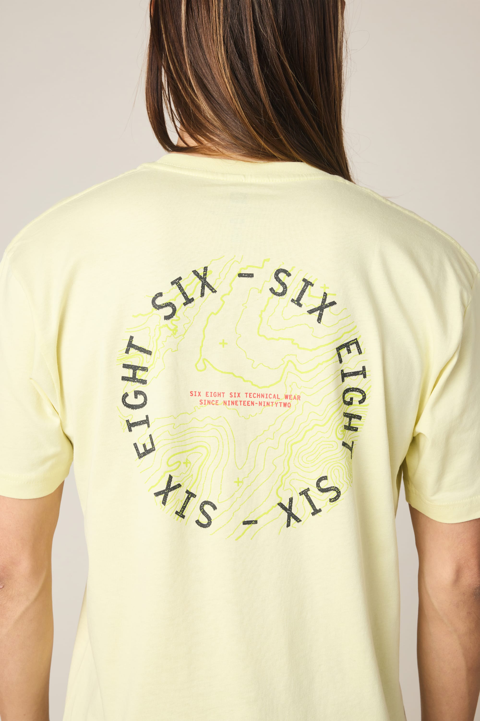 A person wearing the 686 Lifts Authentic SS Tee, a men's 100% cotton t-shirt in light yellow by 686, with "SIX EIGHT SIX" and "TECHNICAL TEAM SINCE NINETEEN NINETY TWO" printed on the back in a circle over a contour map design.