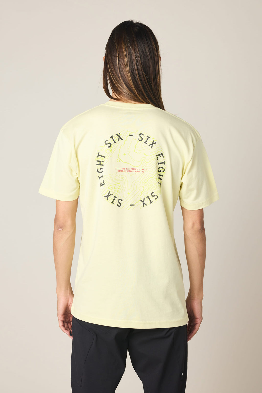Person with long hair, wearing a pale yellow 686 Lifts Authentic SS Tee (100% cotton) and black pants, facing away. The back of the 686 tee shows a circular SIX EIGHT graphic with stylized lines. Neutral background.
