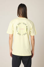 Person with long hair, wearing a pale yellow 686 Lifts Authentic SS Tee (100% cotton) and black pants, facing away. The back of the 686 tee shows a circular SIX EIGHT graphic with stylized lines. Neutral background.