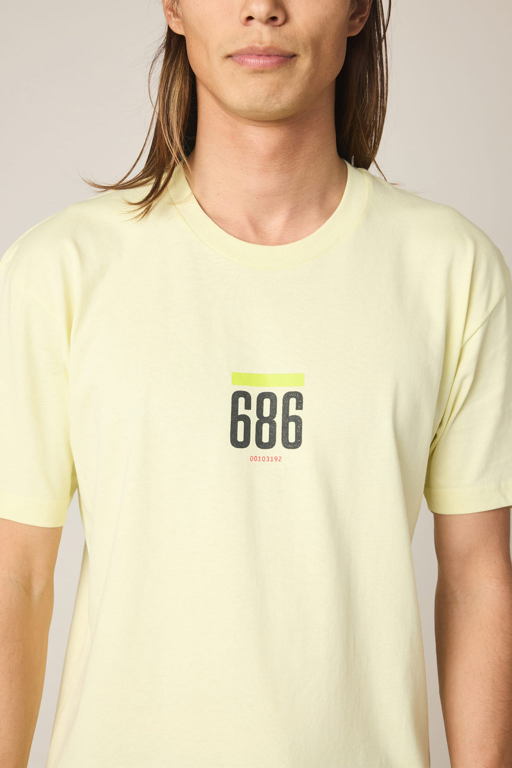 A person with long brown hair (eyes not visible) wears the 686 Lifts Authentic SS Tee by 686, a light yellow 100% cotton men's shirt featuring "686" in black on the chest, a neon green line above, and small red text below.