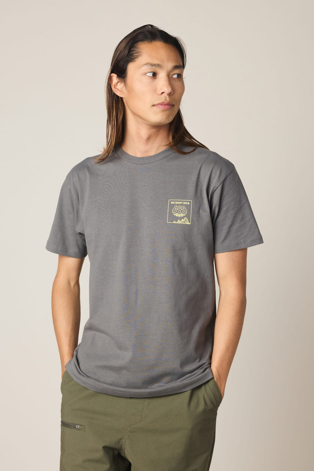 A person with long brown hair wears the 686 Cross-Cut Authentic S/S Tee in gray with a small yellow graphic on the left chest, paired with olive green pants, standing hands in pockets and looking to the side against a plain background.