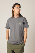 A person with long brown hair wears the 686 Cross-Cut Authentic S/S Tee in gray with a small yellow graphic on the left chest, paired with olive green pants, standing hands in pockets and looking to the side against a plain background.