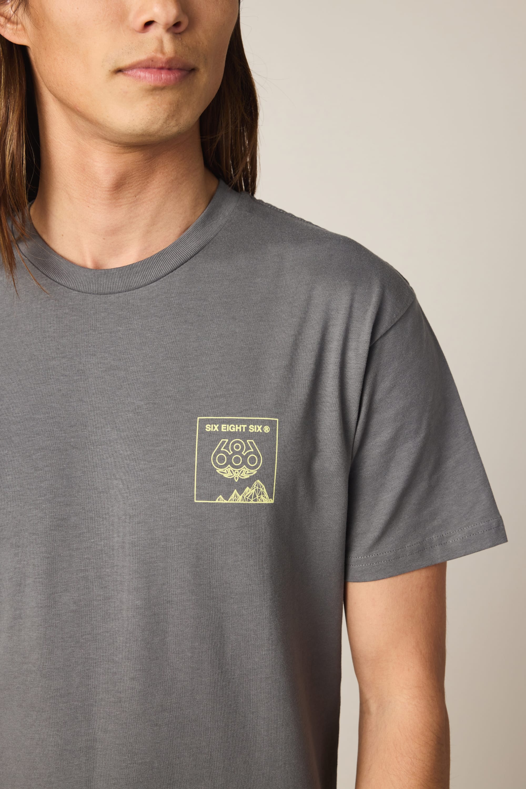 A person with long hair wears the 686 Cross-Cut Authentic S/S Tee by 686 in grey, featuring a small yellow graphic with "SIX EIGHT SIX," "686," and a mountain design on the left chest.