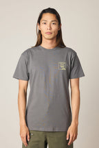 A person with long brown hair wears a gray 686 Cross-Cut Authentic S/S Tee by 686, featuring a small yellow graphic on the left chest, while standing and facing the camera against a plain, light background.