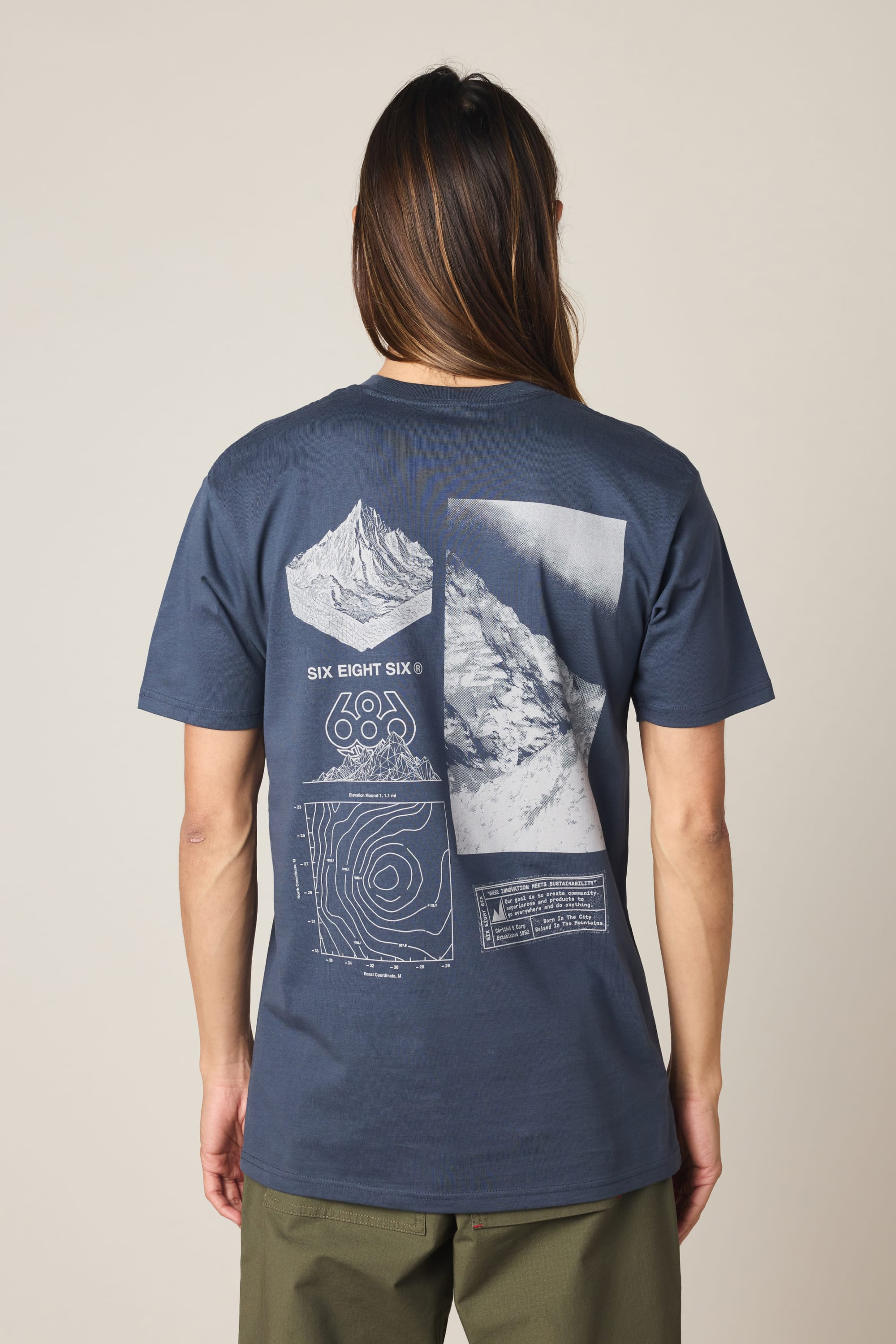 A person with long hair, seen from behind, wears the 686 Cross-Cut Authentic S/S Tee in navy by 686, featuring mountain graphics, contour lines, "686" numbers, and back text. The outfit is paired with olive green pants.