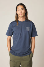 A person with long hair wears the 686 Cross-Cut Authentic S/S Tee by 686 in navy blue with a small white chest graphic and olive green pants, standing against a plain light background.