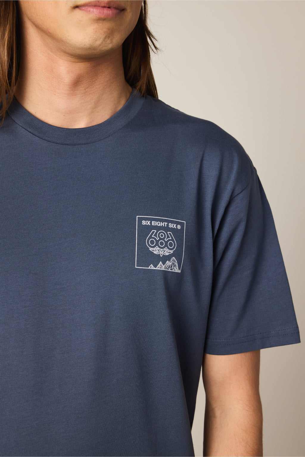 A person wears the 686 Cross-Cut Authentic S/S Tee, a blue men's cotton t-shirt by 686 with a small chest logo displaying "SIX EIGHT SIX," geometric shapes, and mountain outlines. Only their lower face, neck, and upper torso are visible.