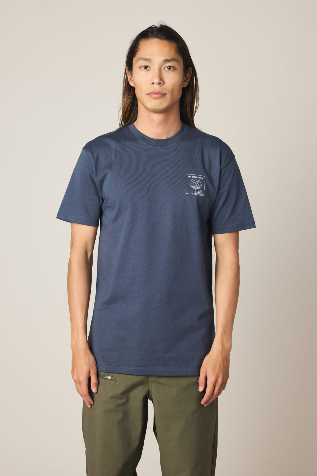Wearing the 686 Cross-Cut Authentic S/S Tee in navy blue with a small white chest graphic, paired with olive green pants, a long-haired person stands against a plain light background.