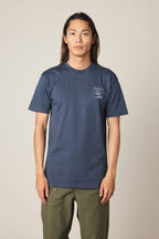 Wearing the 686 Cross-Cut Authentic S/S Tee in navy blue with a small white chest graphic, paired with olive green pants, a long-haired person stands against a plain light background.