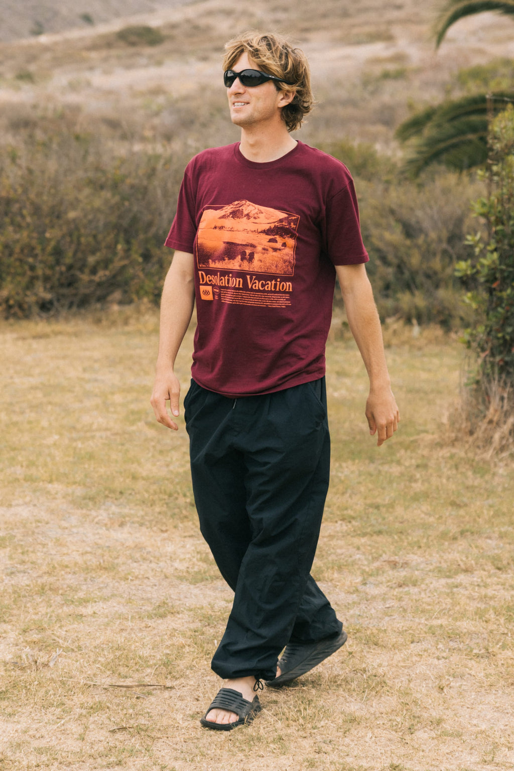 A man wearing sunglasses, black pants, sandals, and the 686 Desolation Vacation Authentic S/S Tee by 686 walks outdoors on dry grass with bushes and a blurred background.