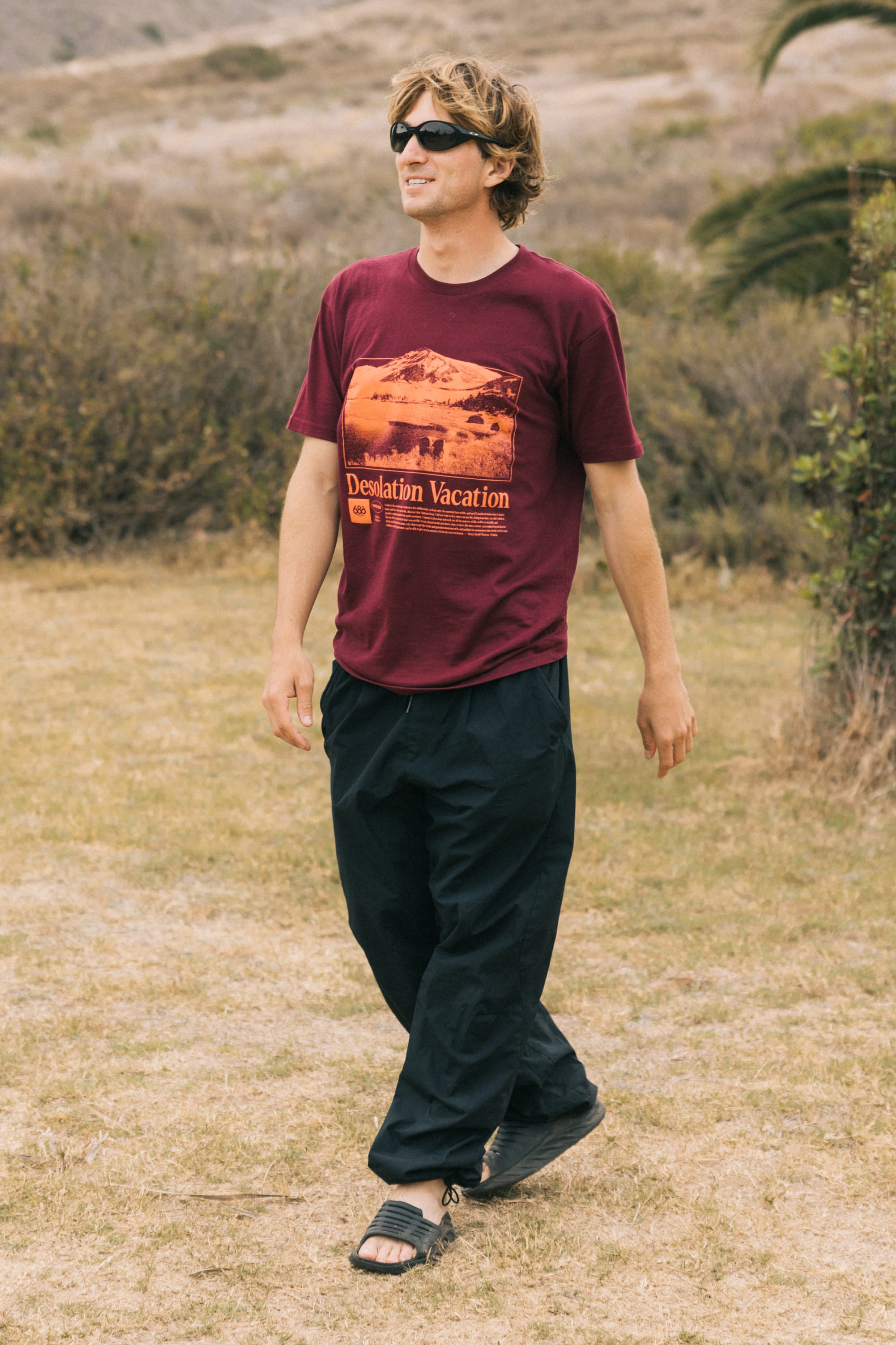 A man wearing sunglasses, black pants, sandals, and the 686 Desolation Vacation Authentic S/S Tee by 686 walks outdoors on dry grass with bushes and a blurred background.