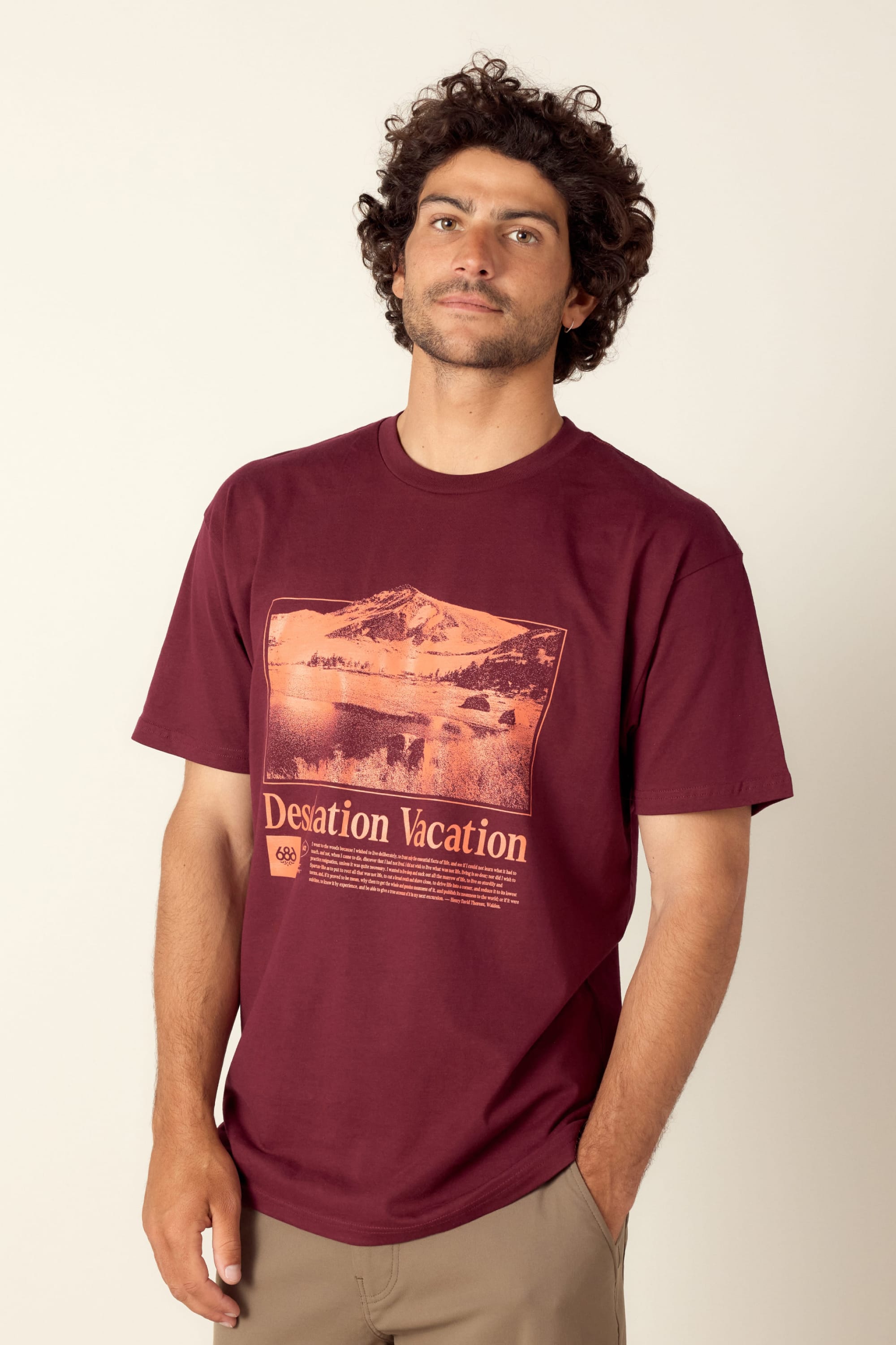 A man with curly hair wears the 686 Desolation Vacation Authentic S/S Tee, a maroon 100% cotton men’s T-shirt by 686 with an orange mountain landscape and Desolation Vacation text. He stands against a neutral background, looking at the camera.
