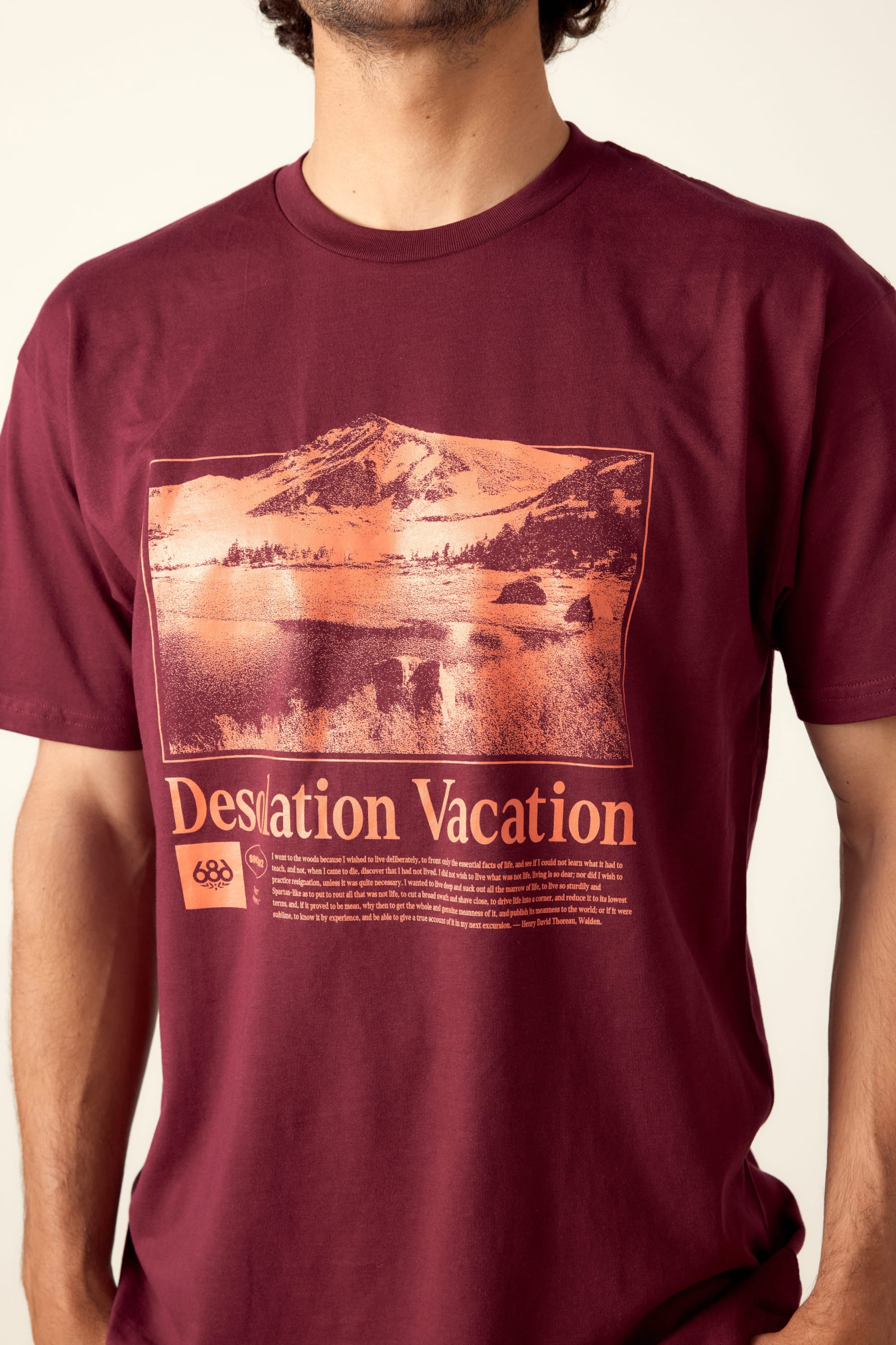 A person wearing the 686 Desolation Vacation Authentic S/S Tee by 686, a maroon mens 100% cotton T-shirt with an orange-tinted mountain, tree, and lake graphic and “Desolation Vacation” printed below the image.