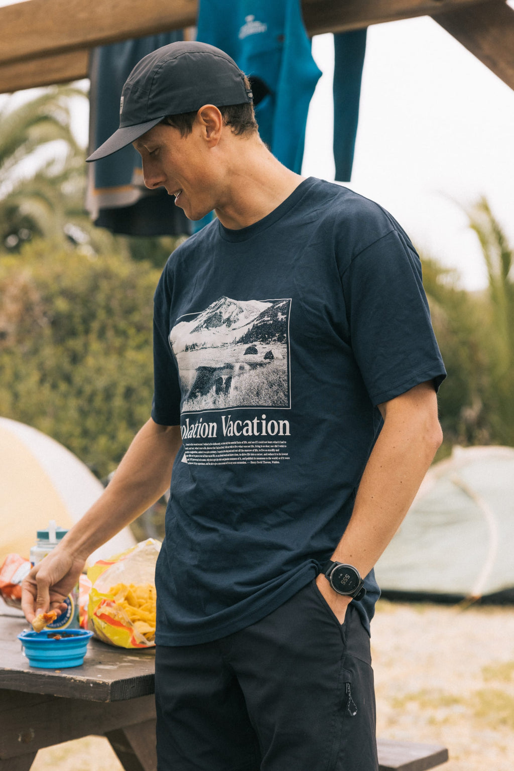 A man in a 686 Desolation Vacation Authentic S/S Tee and navy cap stands outdoors by a snack table, smiling and reaching for a bowl, with tents and greenery visible behind him.