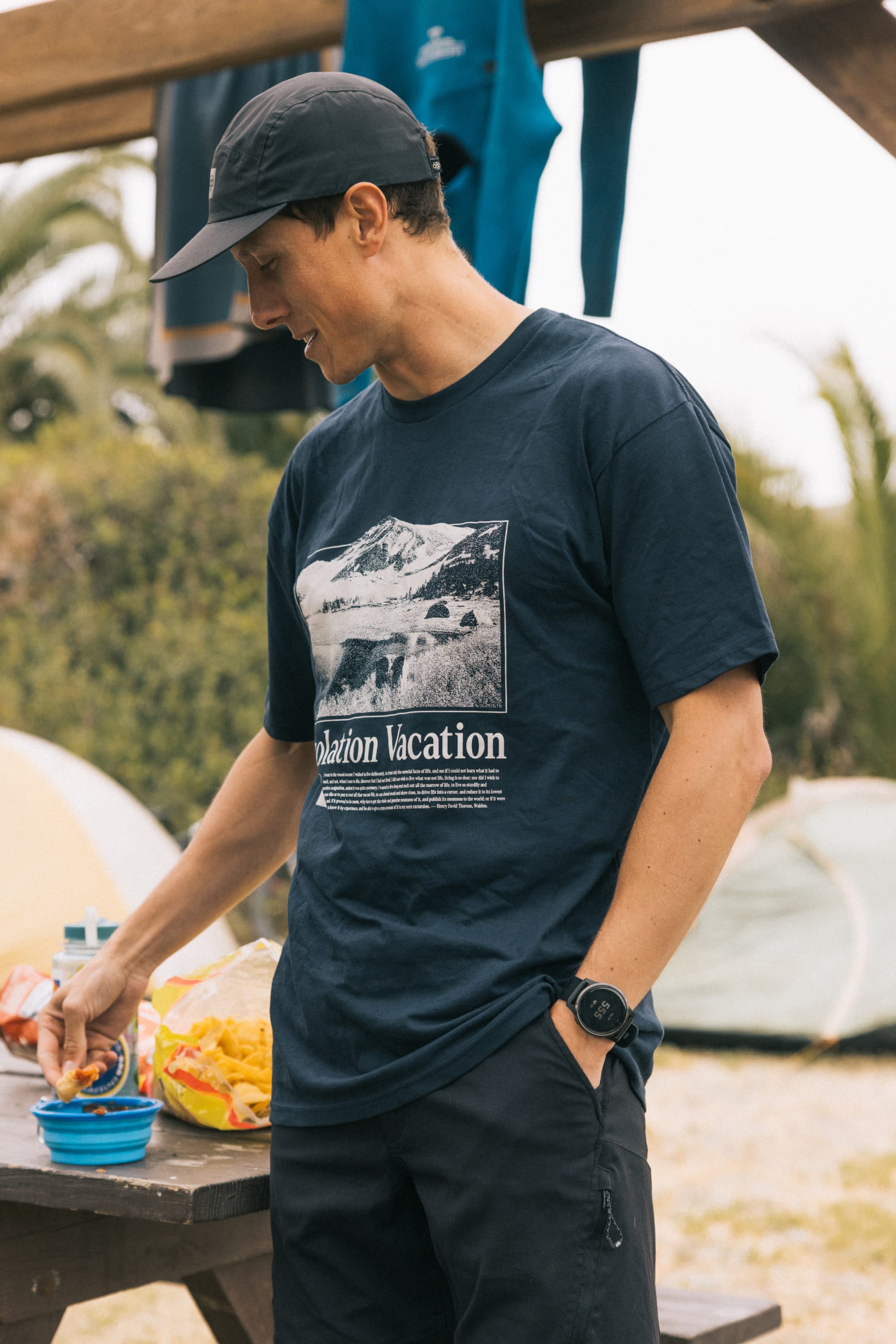 A man in a 686 Desolation Vacation Authentic S/S Tee and navy cap stands outdoors by a snack table, smiling and reaching for a bowl, with tents and greenery visible behind him.
