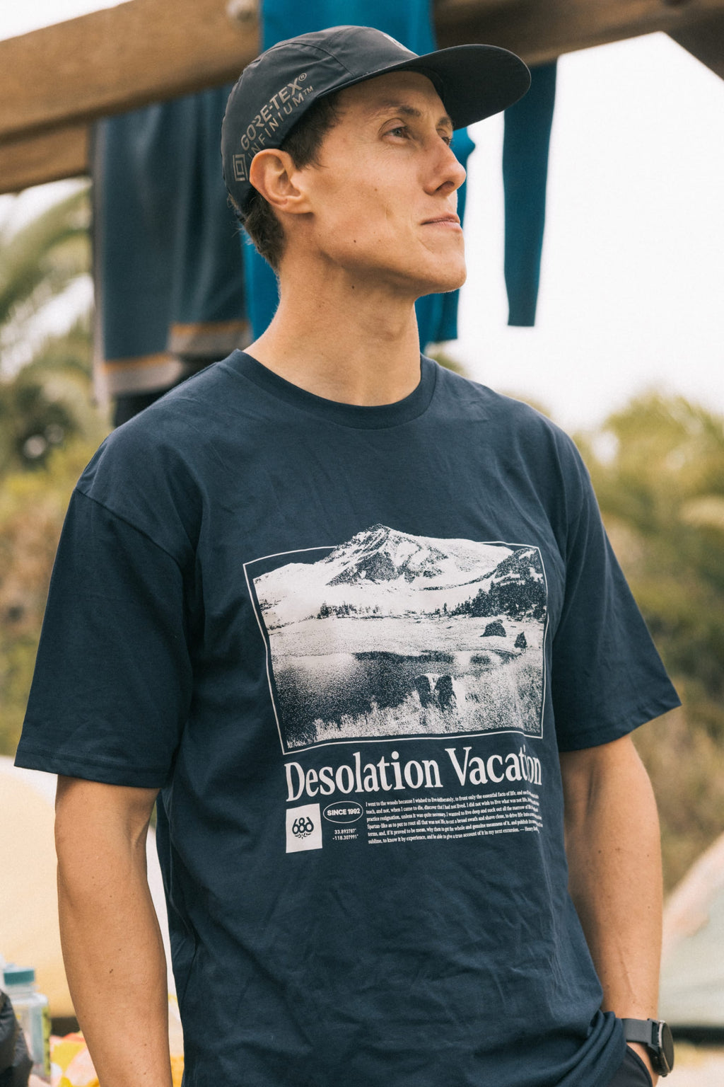 A man outdoors in a dark cap wears the navy blue 686 Desolation Vacation Authentic S/S Tee by 686, made from 100% cotton with a mountain and lake graphic and “Desolation Vacation” text. Trees and hanging clothes are visible in the background.