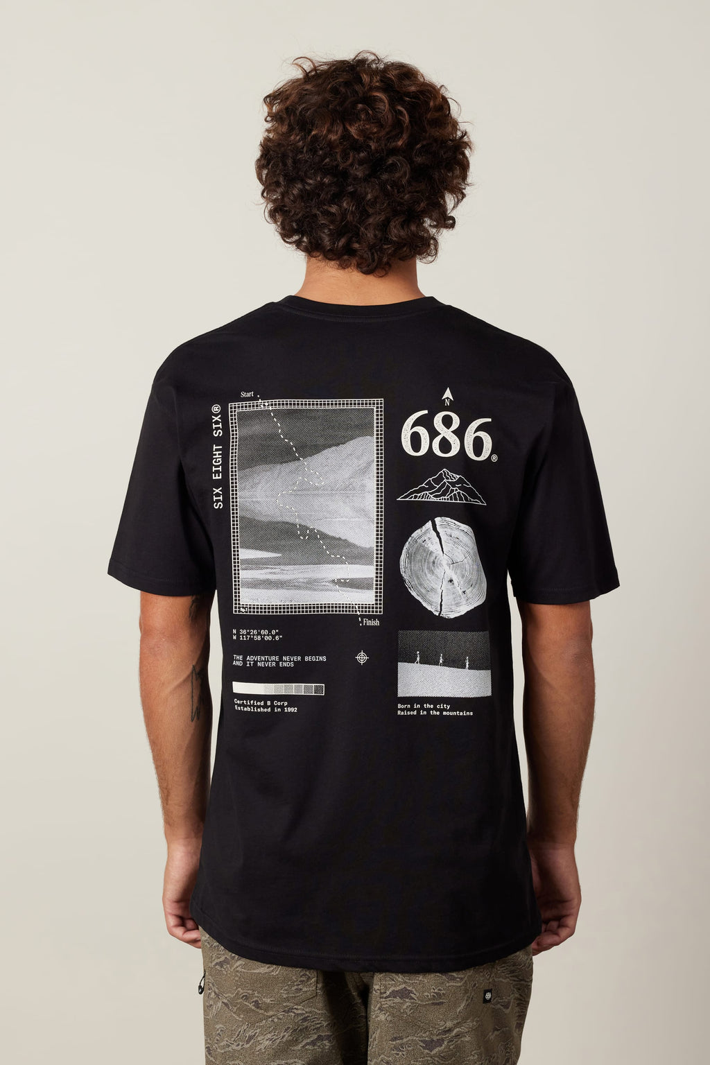 A person with curly hair wears the 686 Path Authentic S/S Tee by 686, a 100% cotton men’s black T-shirt featuring white scientific graphics, geometric shapes, numbers, and diagrams on the back. The shirt is shown from behind.