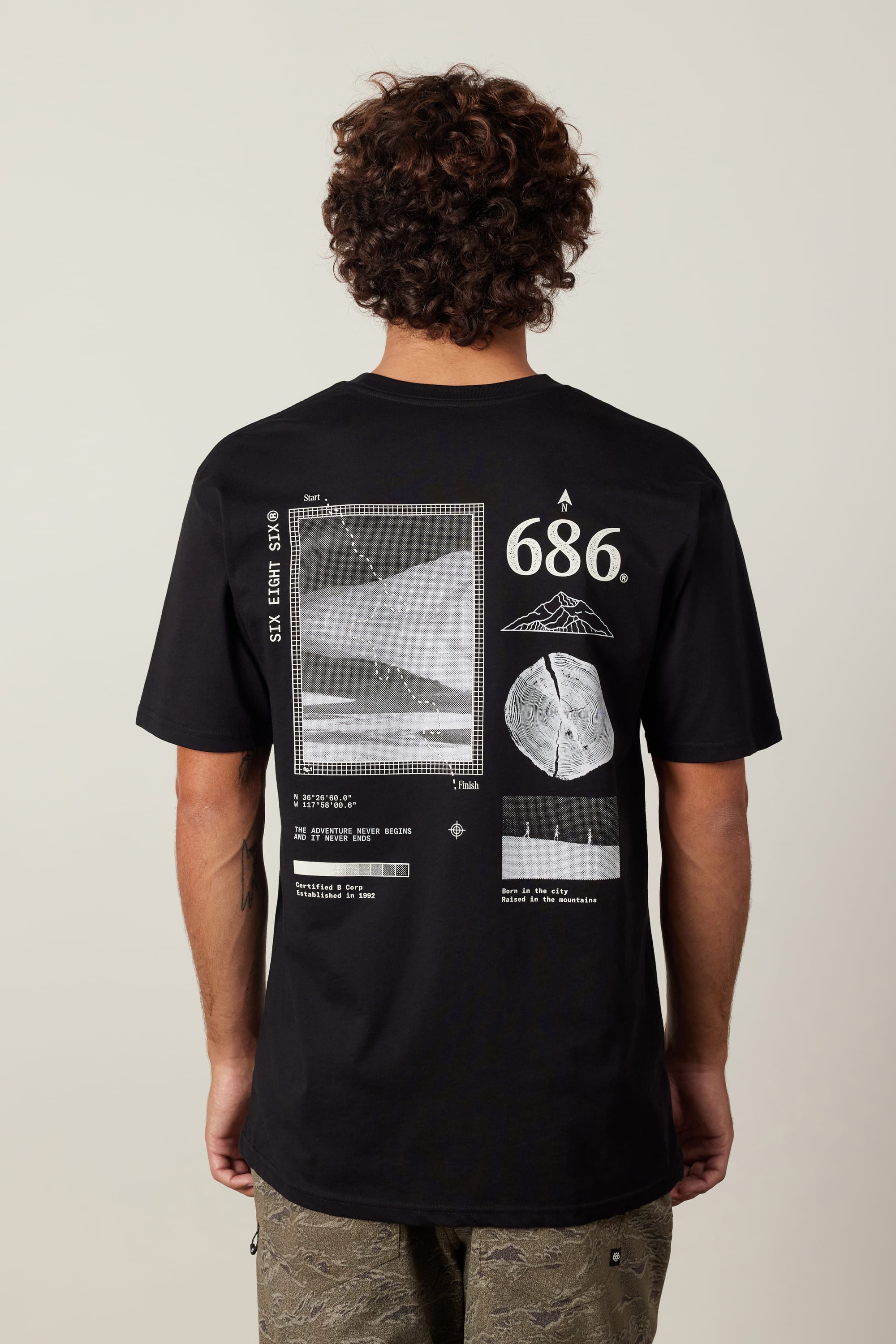 A person with curly hair wears the 686 Path Authentic S/S Tee by 686, a 100% cotton men’s black T-shirt featuring white scientific graphics, geometric shapes, numbers, and diagrams on the back. The shirt is shown from behind.