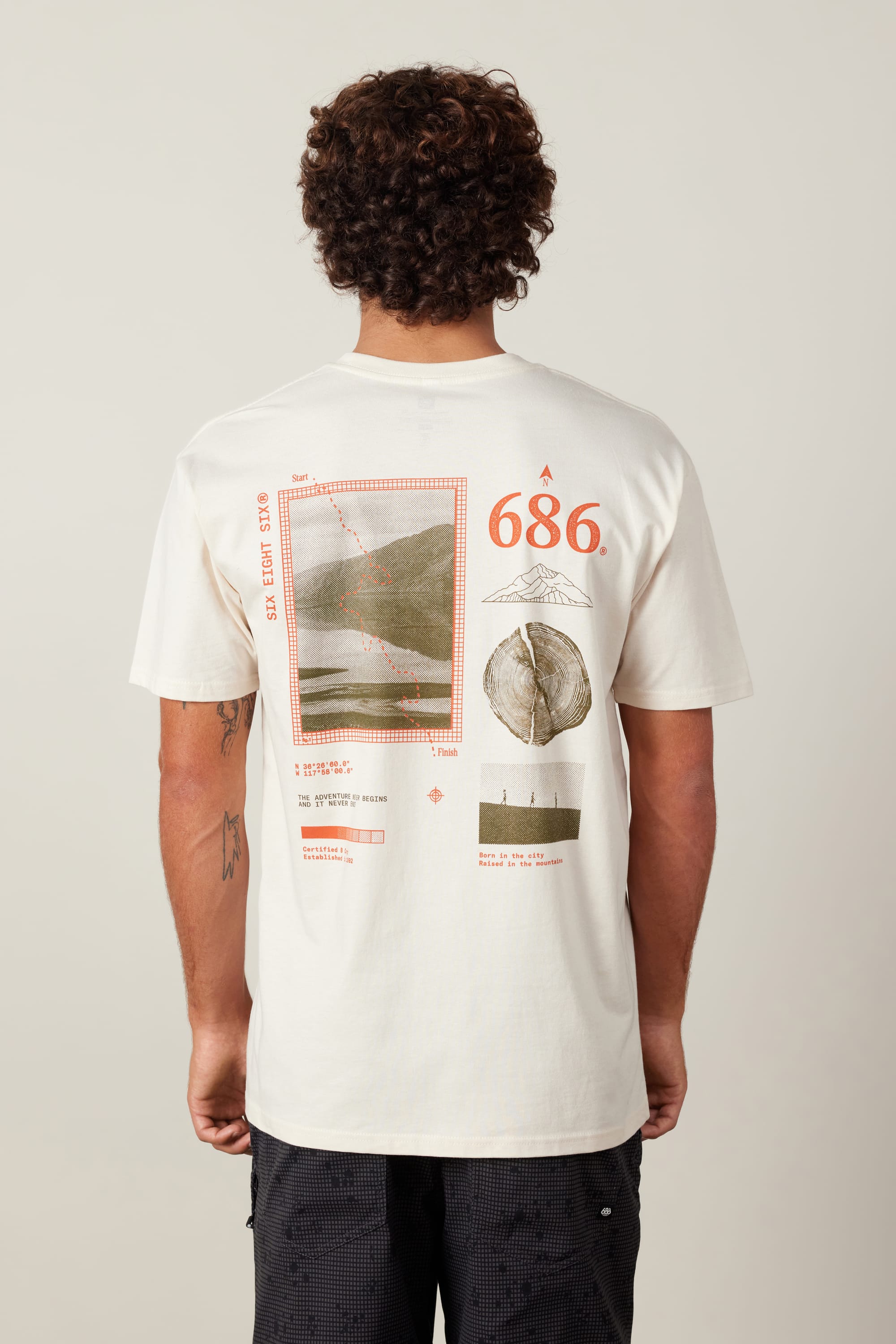 A person with curly hair is seen from behind wearing the 686 Path Authentic S/S Tee by 686, a white 100% cotton men's t-shirt with abstract graphics, red "686" text, and geometric designs on the back.