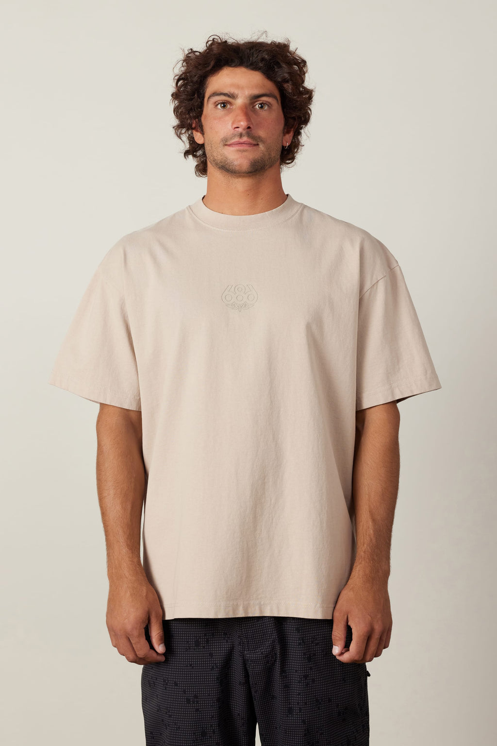 A man with curly brown hair wears the 686 High Def Relaxed S/S Tee by 686 in beige, paired with dark patterned shorts, standing facing forward against a plain light background.