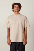 A man with curly brown hair wears the 686 High Def Relaxed S/S Tee by 686 in beige, paired with dark patterned shorts, standing facing forward against a plain light background.