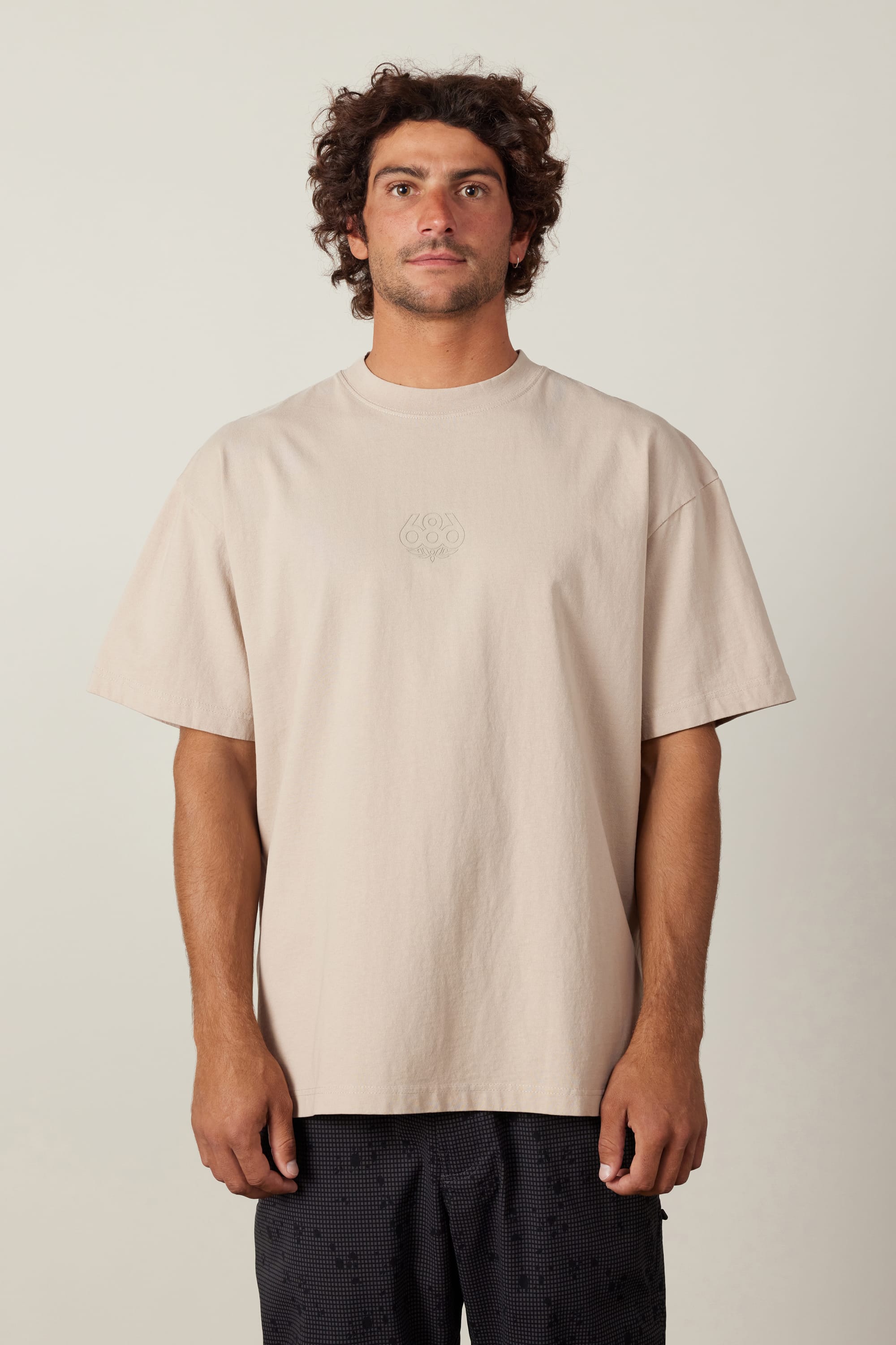 A man with curly brown hair wears the 686 High Def Relaxed S/S Tee by 686 in beige, paired with dark patterned shorts, standing facing forward against a plain light background.