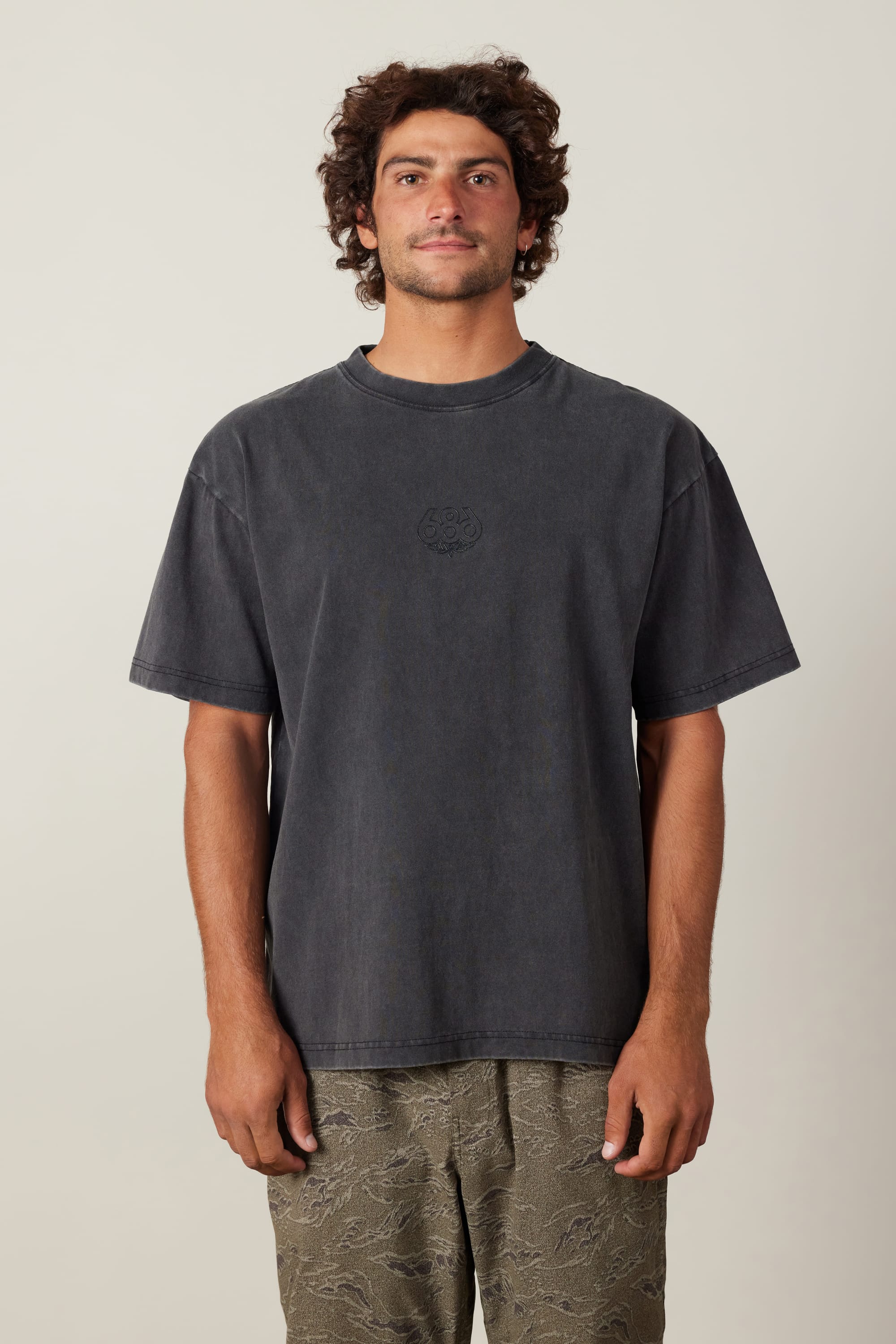 A man with curly hair stands facing forward against a plain light background, smiling slightly and looking at the camera while wearing the 686 High Def Relaxed S/S Tee by 686 and patterned gray shorts.