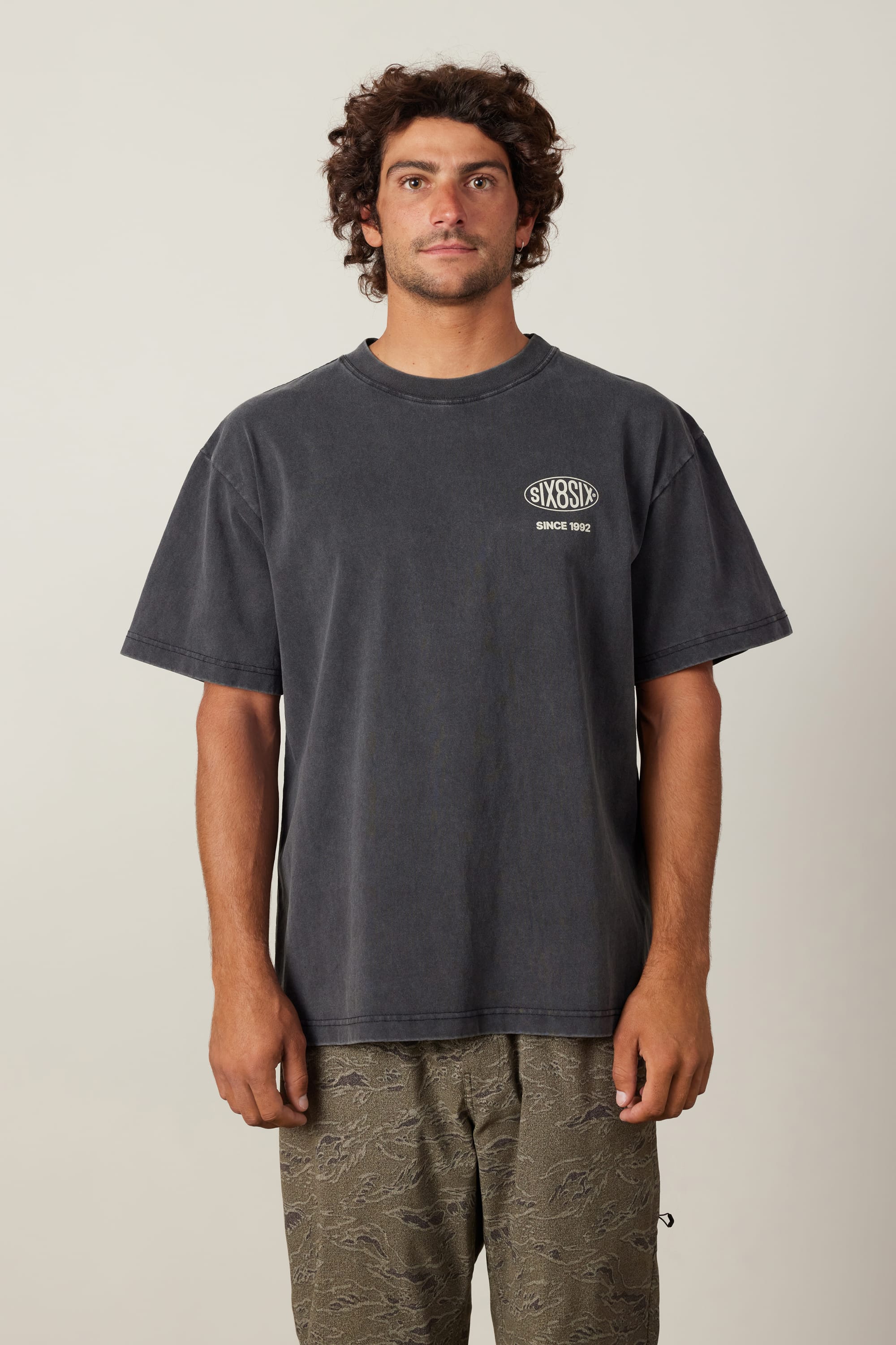 A man with curly hair and a mustache wears the 686 92 Relaxed S/S Tee by 686—a loose, faded black 100% cotton t-shirt with a white logo—paired with patterned gray-green shorts, standing against a plain light background.