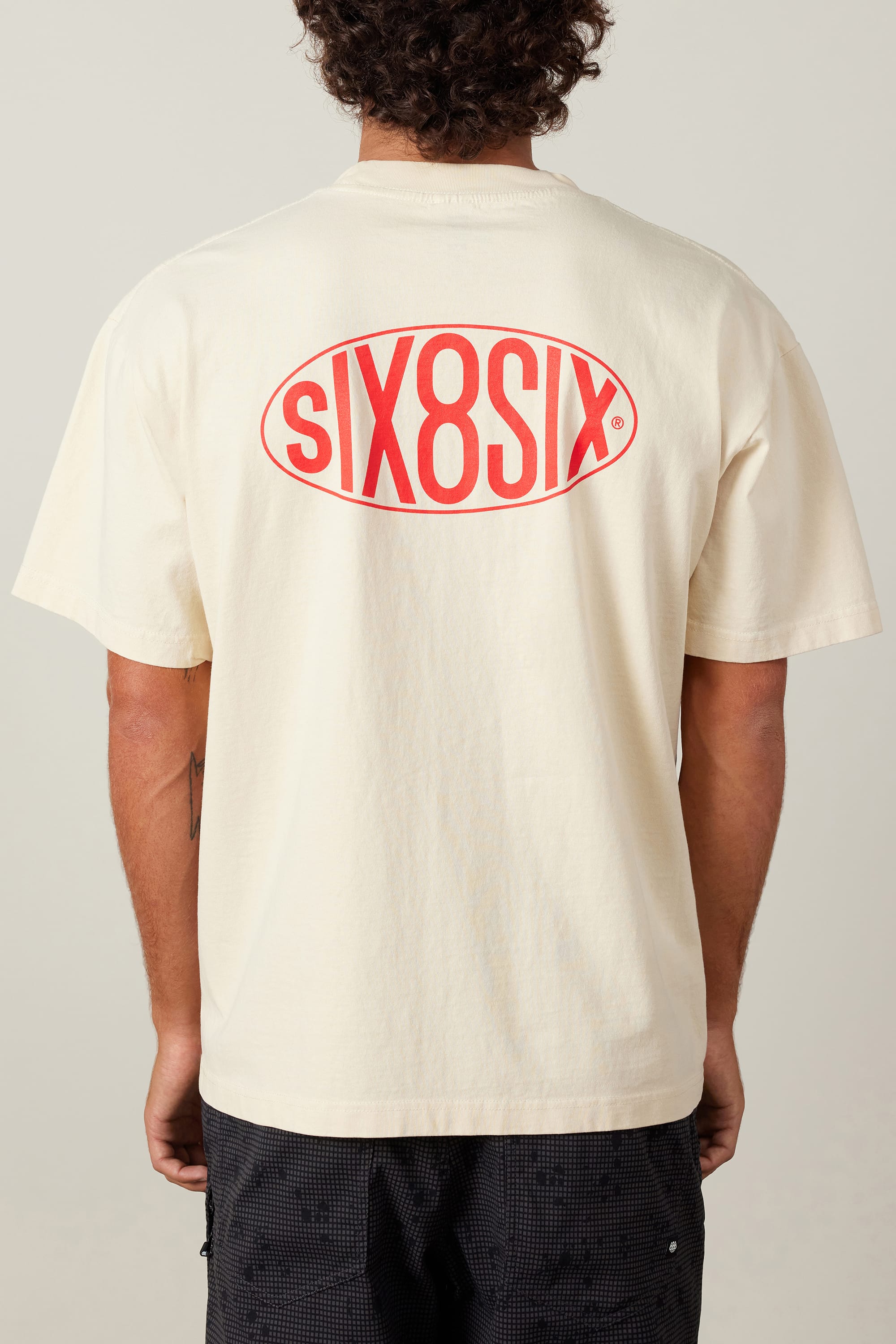 A person stands facing away, wearing a light beige 686 92 Relaxed S/S Tee by 686, made from 100% cotton, featuring a large red oval siX8siX logo in bold letters on the back.
