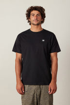 A man with curly brown hair wears the 686 Logo 3-Pack S/S Tee by 686, a plain black 100% cotton men's t-shirt with a small white logo on the left chest, paired with patterned green shorts against a light background.