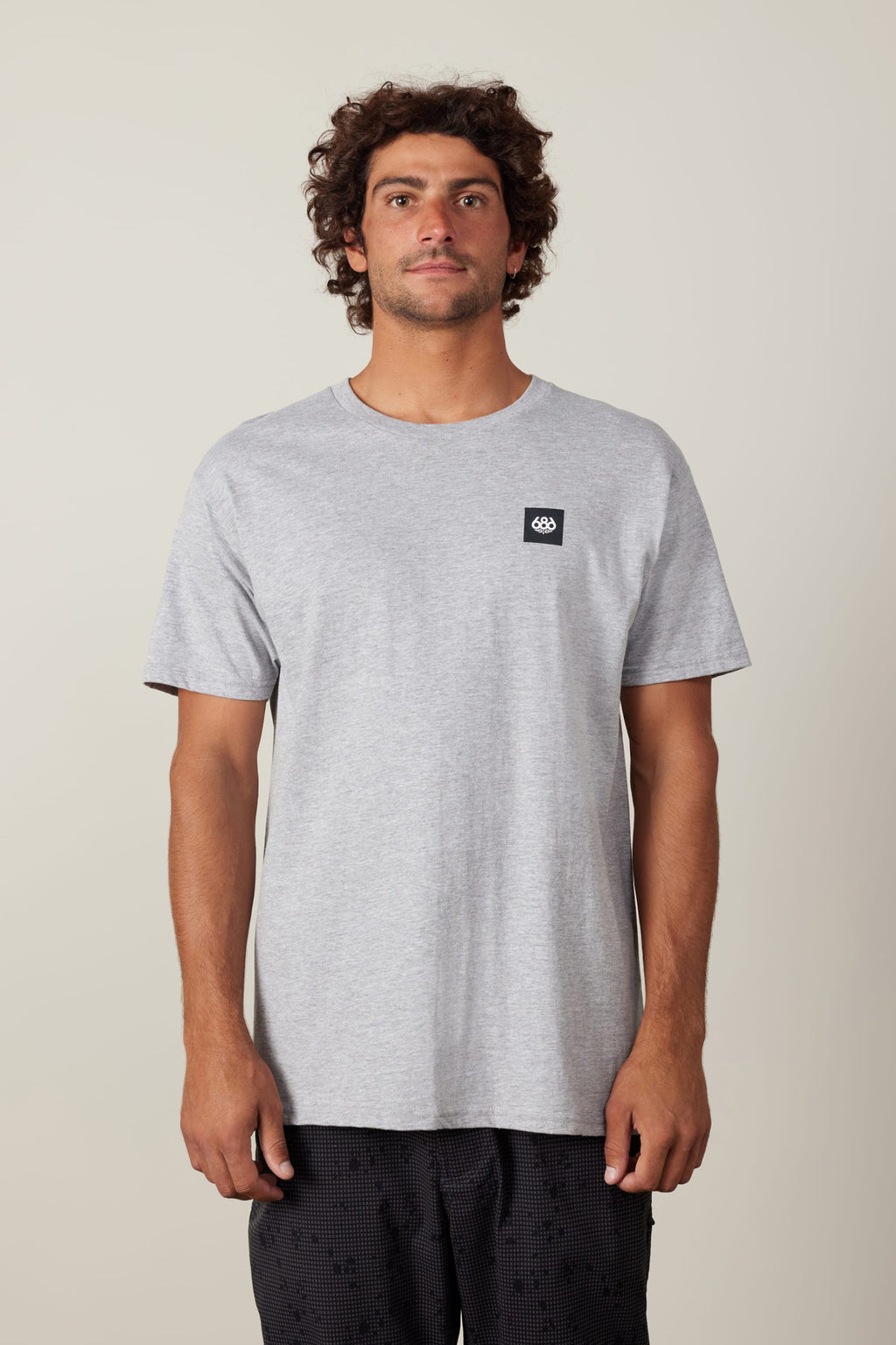 A man with curly dark hair stands against a plain light background, wearing the 686 Logo 3-Pack S/S Tee in light gray with a small black logo on the left chest and black patterned shorts, facing forward with a neutral expression.