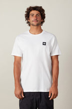 A man with curly brown hair wears a plain white 686 Logo 3-Pack S/S Tee by 686 featuring a small black logo on the left chest and black patterned shorts, standing against a light background.