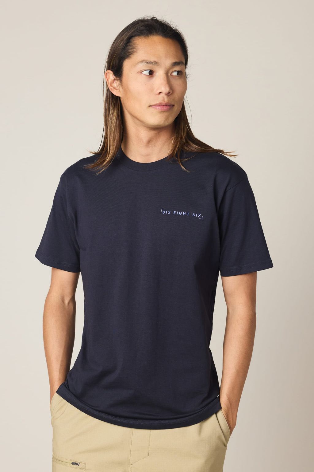 A person with long brown hair wears the 686 24/7 Authentic SS Tee by 686—a navy blue men’s t-shirt with “SIX EIGHT SIX” on the chest—paired with beige pants, hands in pockets, looking sideways against a plain background. Made from 100% cotton.