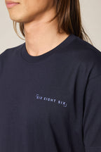 A person with long brown hair wears a 686 24/7 Authentic SS Tee—a dark navy men's t-shirt made from 100% cotton with "SIX EIGHT SIX" in white on the chest. The image shows the upper torso and part of their face.