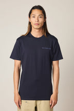 A person with long brown hair stands facing forward against a plain light background, wearing the 686 24/7 Authentic SS Tee by 686—a navy blue men's t-shirt made from 100% cotton with small white text on the left chest—and beige pants.