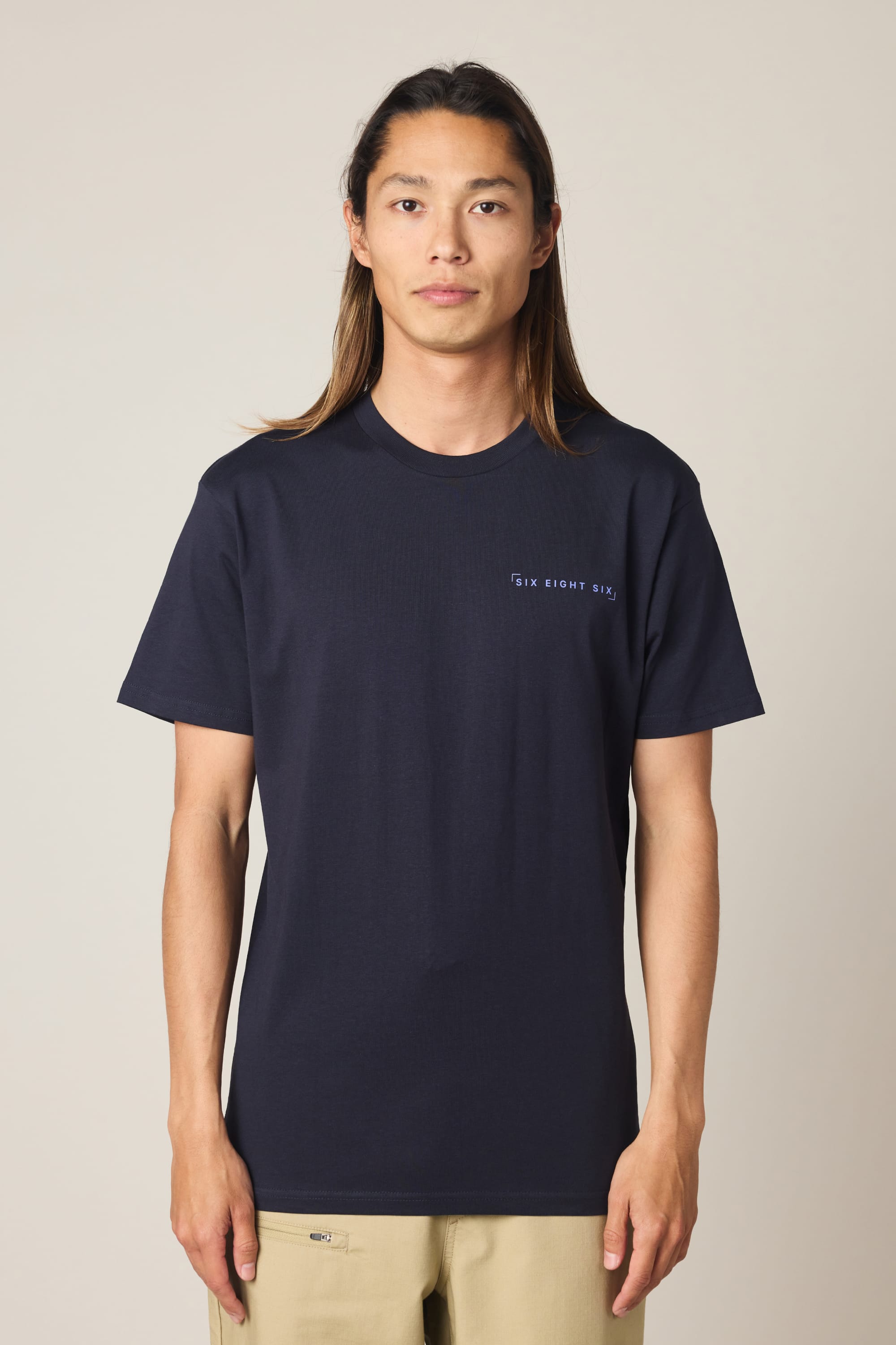 A person with long brown hair stands facing forward against a plain light background, wearing the 686 24/7 Authentic SS Tee by 686—a navy blue men's t-shirt made from 100% cotton with small white text on the left chest—and beige pants.