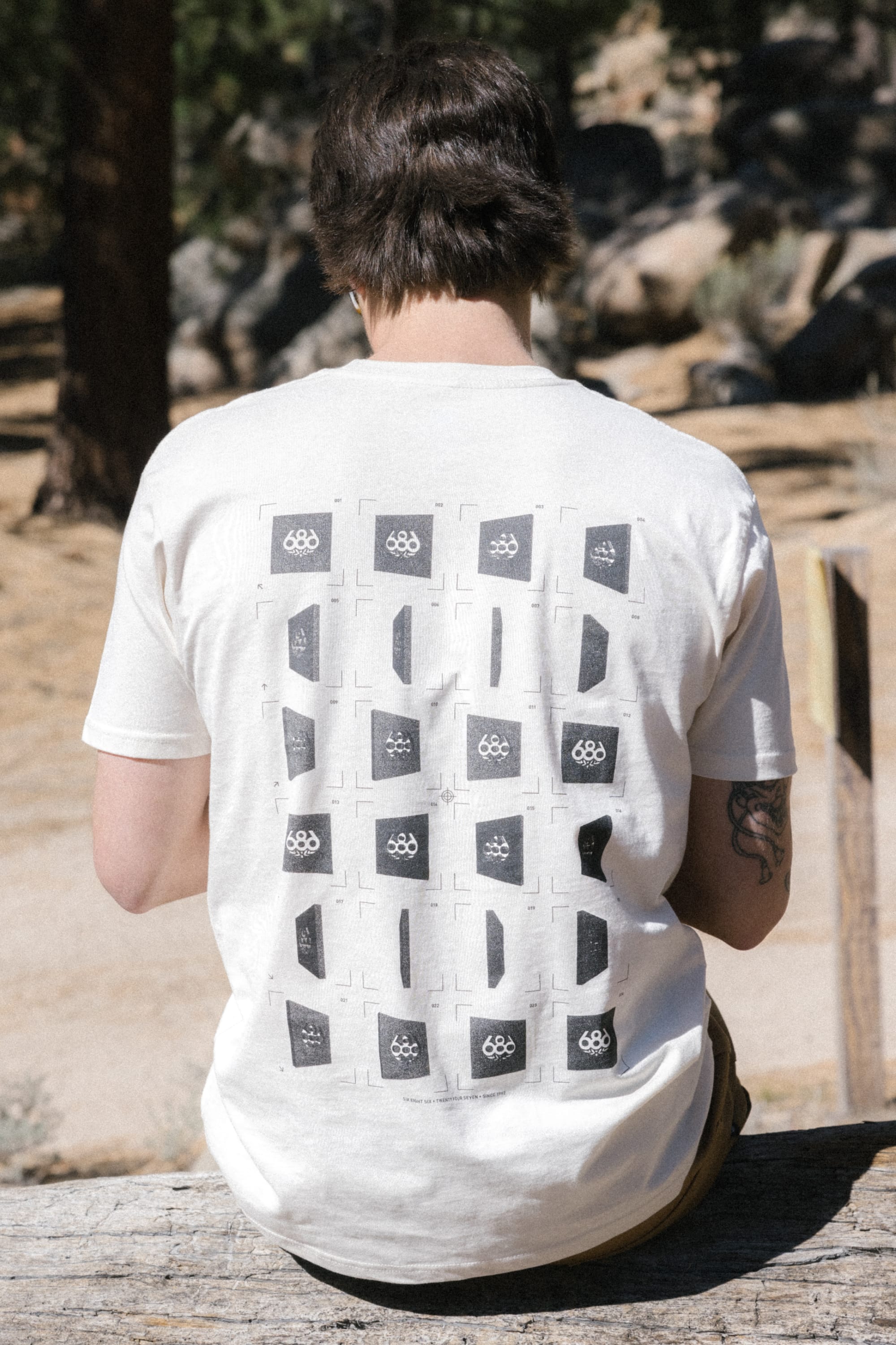 A person with short brown hair sits outdoors on a wooden rail, wearing the 686 24/7 Authentic SS Tee by 686, which features black and white geometric designs and Olympic rings on the back. Trees and rocks are in the background.